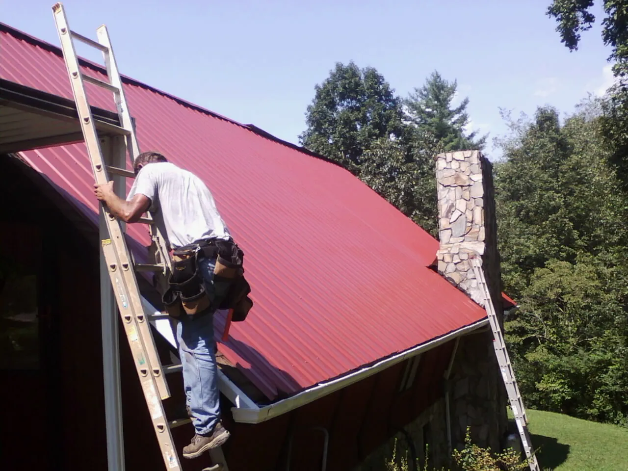 Roofer on a ladder beside a red metal roof and stone chimney — Soffit & Fascia Repair by Roofing Brothers in Equinunk